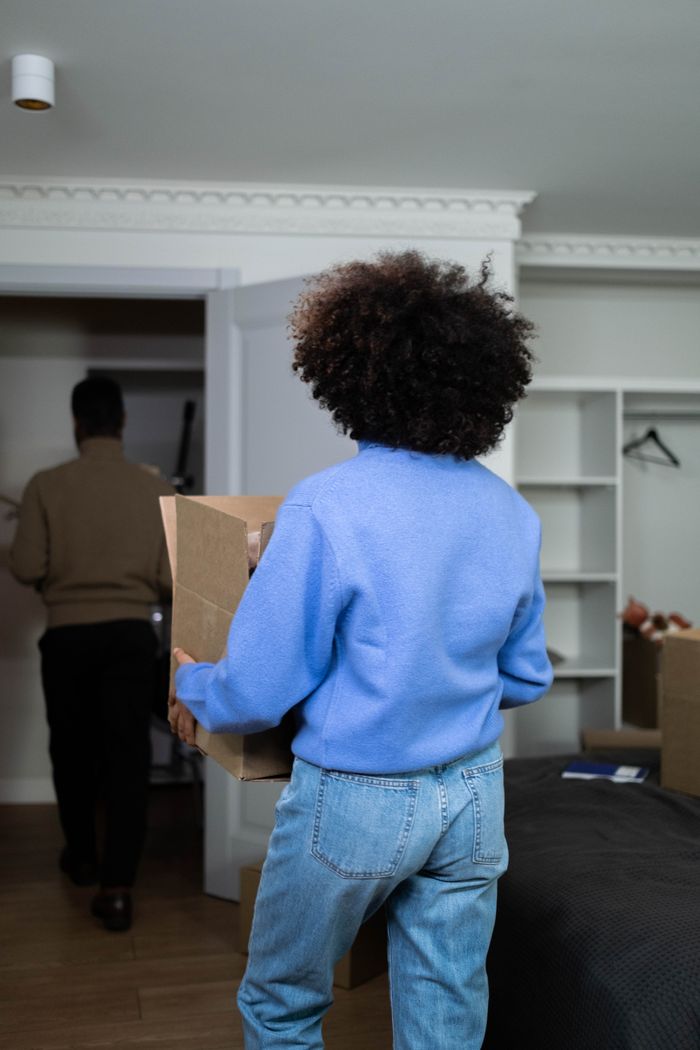 Vue de dos d’une femme portant une boîte à la maison lors du jour d’emménagement, mettant l’accent sur le thème du déménagement et de la relocalisation.